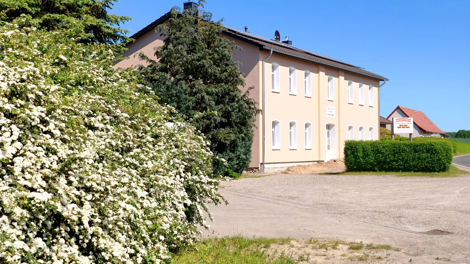 Erbkrug Blankensee -  Ferienapartments - Hinter einem üppig blühenden Busch mit weißen Blüten steht ein großes beigefarbenes zweistöckiges Gebäude mit braunem Dach.