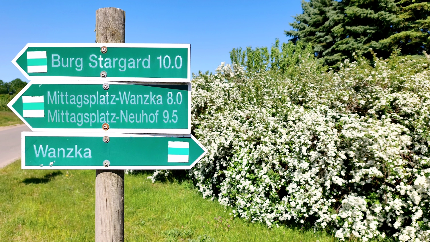 Erbkrug Blankensee -  Ferienapartments - Grüne Wegweiser auf einem Holzpfosten weisen vor dem Hintergrund weißblühender Büsche in Richtung Burg Stargard und Wanzka.